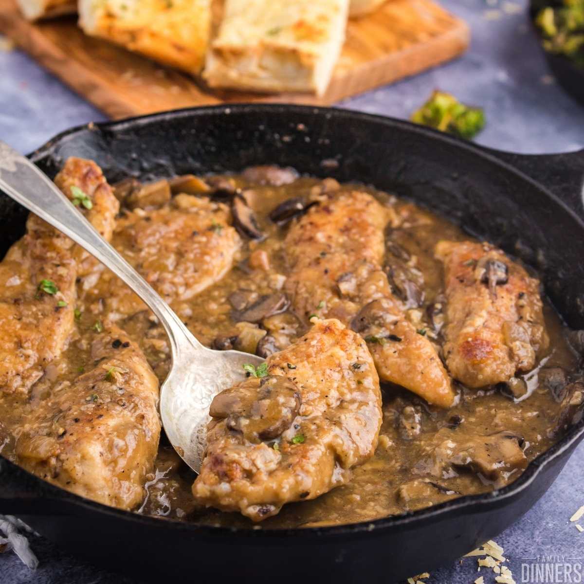 Golden cooked chicken surrounded by mushroom sauce in black cast iron pan. Golden baked french bread slices in background.