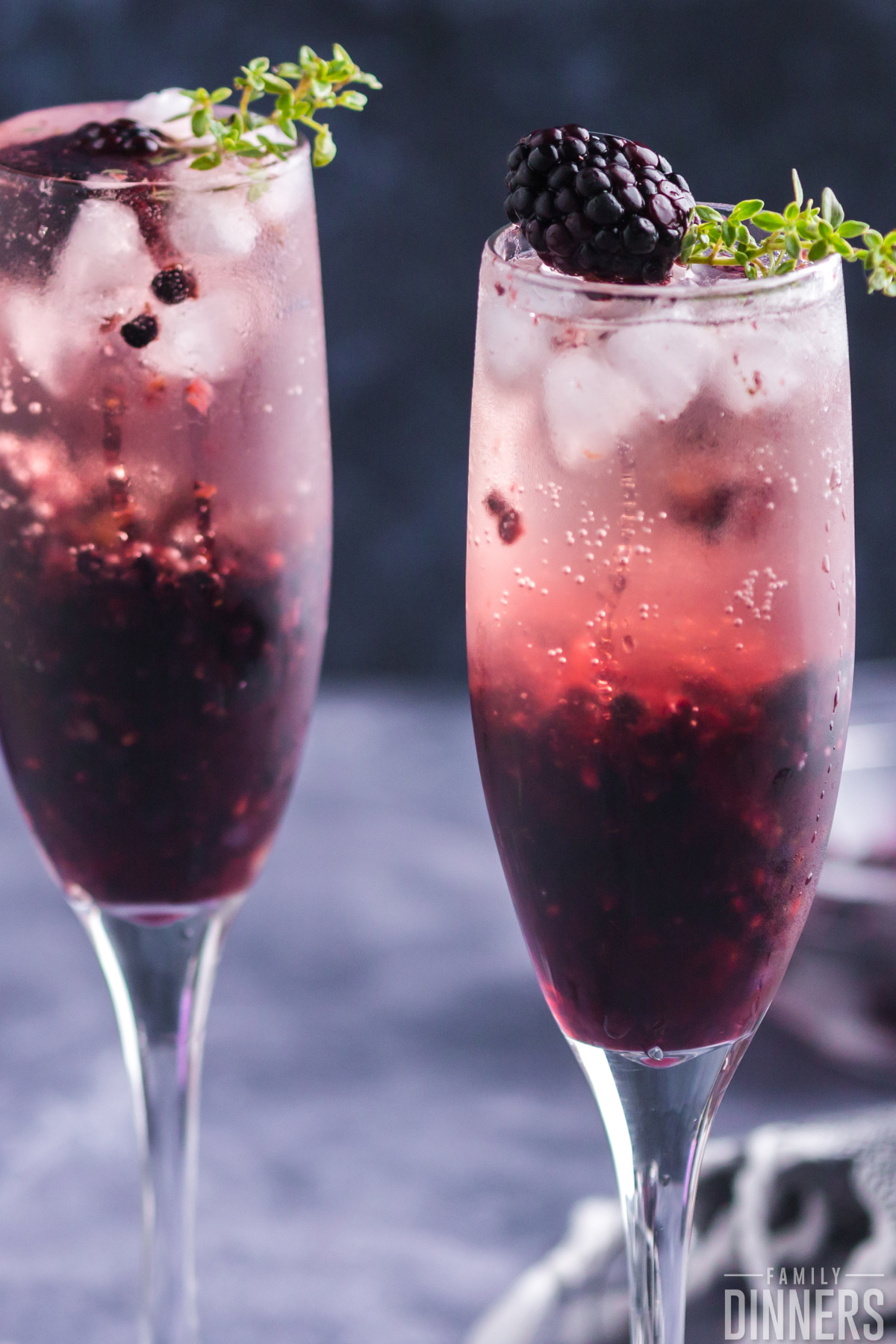 two champagne glasses with blackberry drink inside. Darker blackberry color on the bottom of the glass and light translucent blackberry color at the top. Each glass has fresh thyme and a blackberry on top. Lots of bubbles inside the glass.