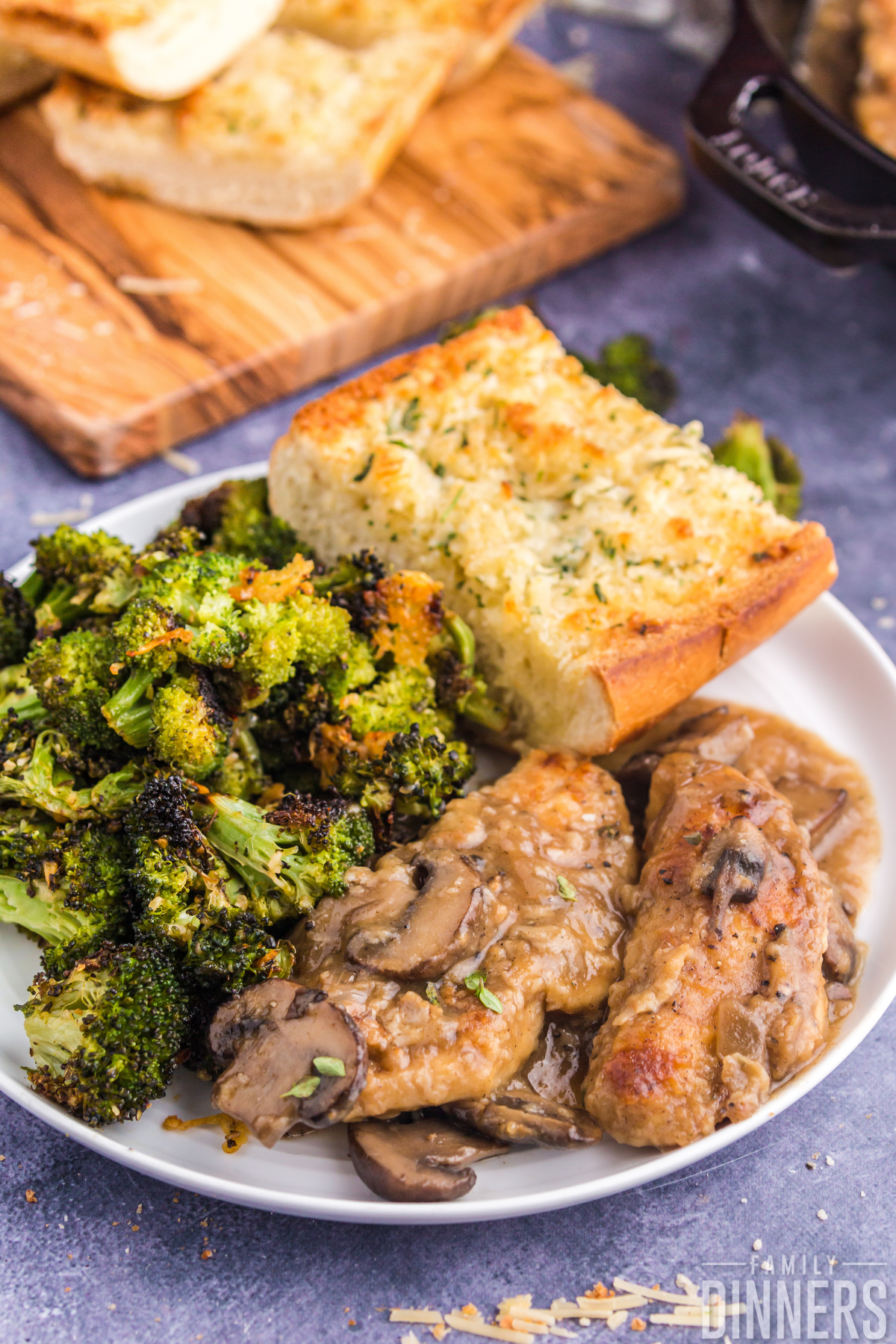 white round plate on blue counter. Plate has two pieces of golden browned chicken with mushrooms and sauce as well as crispy parmesan roasted broccoli and garlic cheese bread slice.