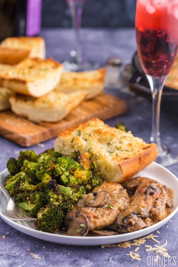 broccoli, french bread, and chicken on white plate, french bread and champagne glass in background