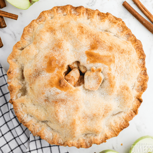 overhead photo of baked apple pie with apple cutout in the top of the crust and cinnamon sticks lying beside