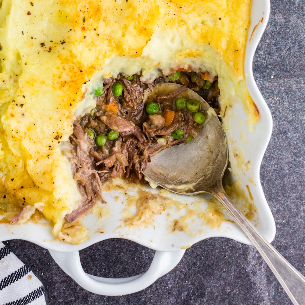 closeup overhead of spoon scooping meat, carrots, peas, and mashed potatoes from white casserole dish