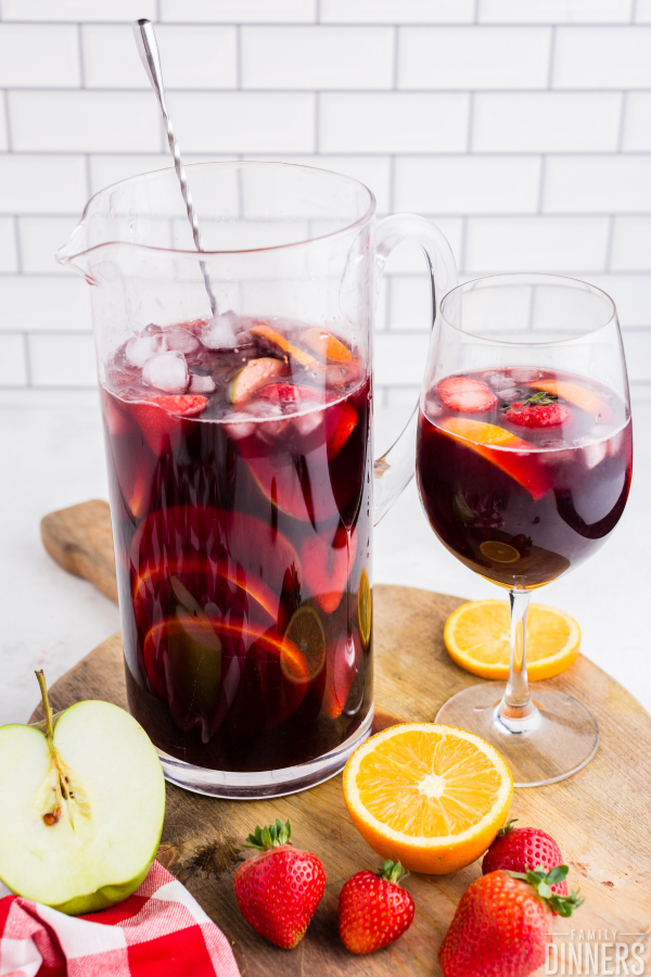 large glass pitcher with silver swizzle stick full of red wine sangria and chopped up fruit next to wine glass full of red wine sangria and fruit. Cut fruit on chopping board.