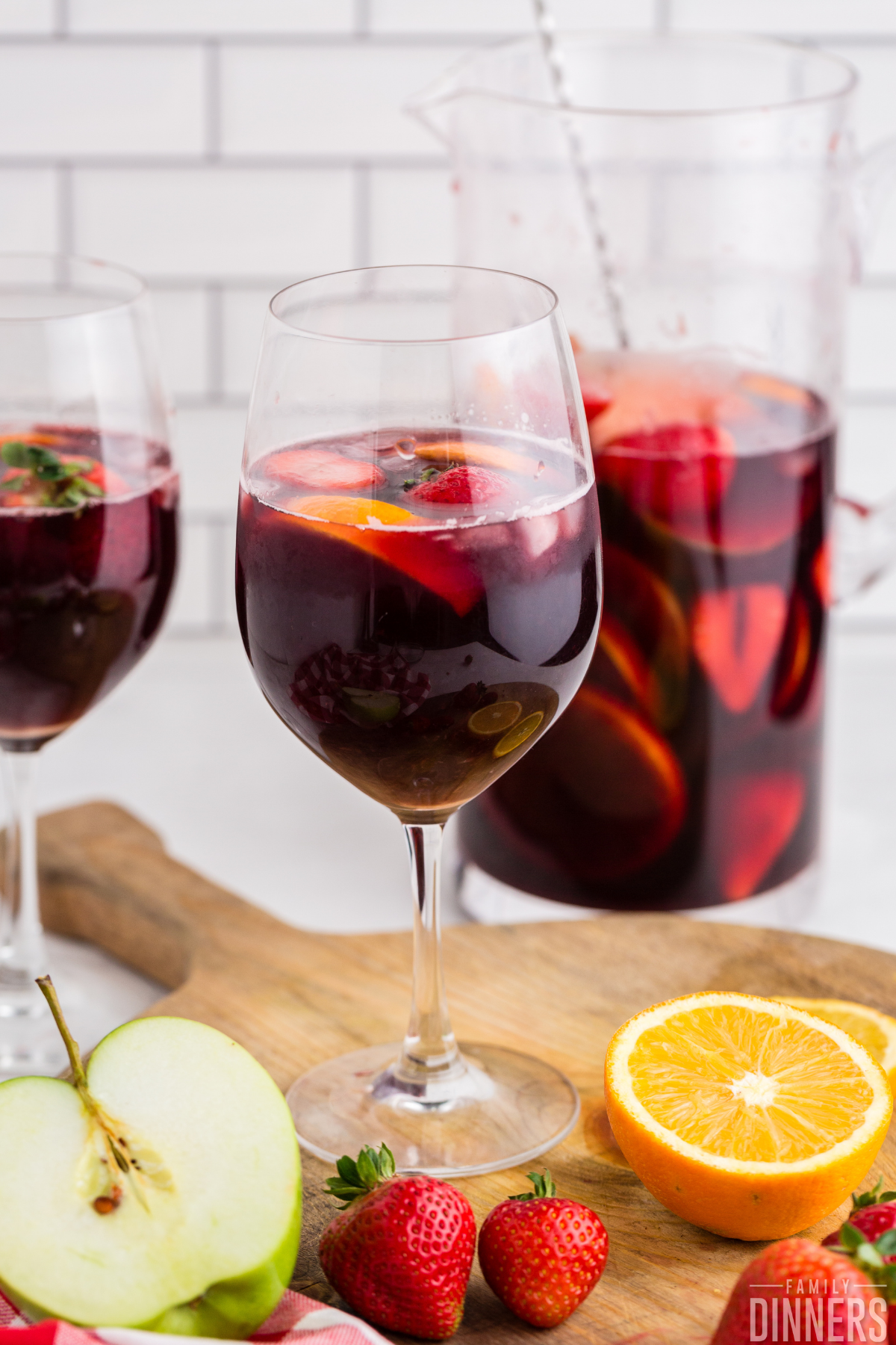 two wine glasses full of red wine sangria with oranges and strawberries and apples floating in them. Glass pitcher in background full of red wine sangria and fruit