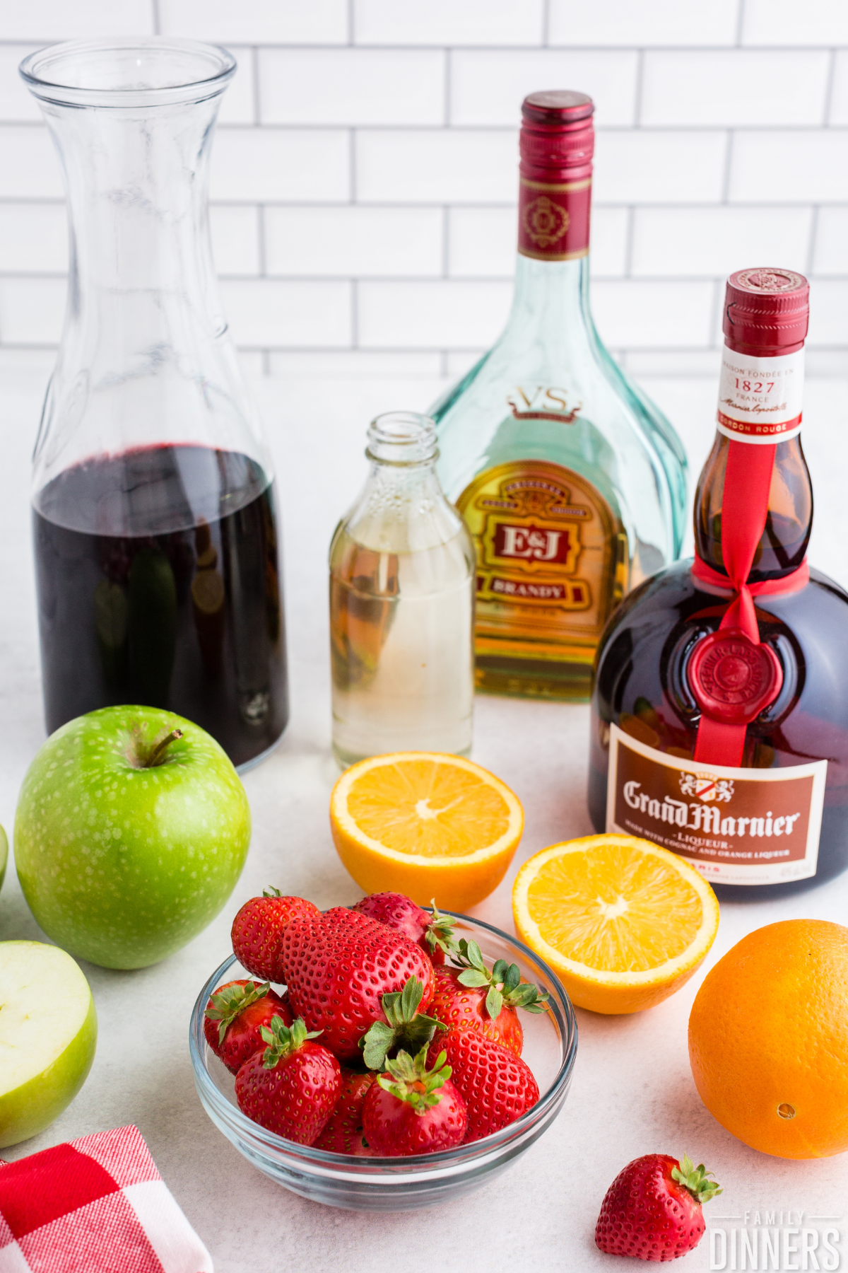 red wine sangria ingredients - grand marnier, brandy, simple syrup, red wine, apple, orange, strawberries on white counter with white subway tile backgroundw