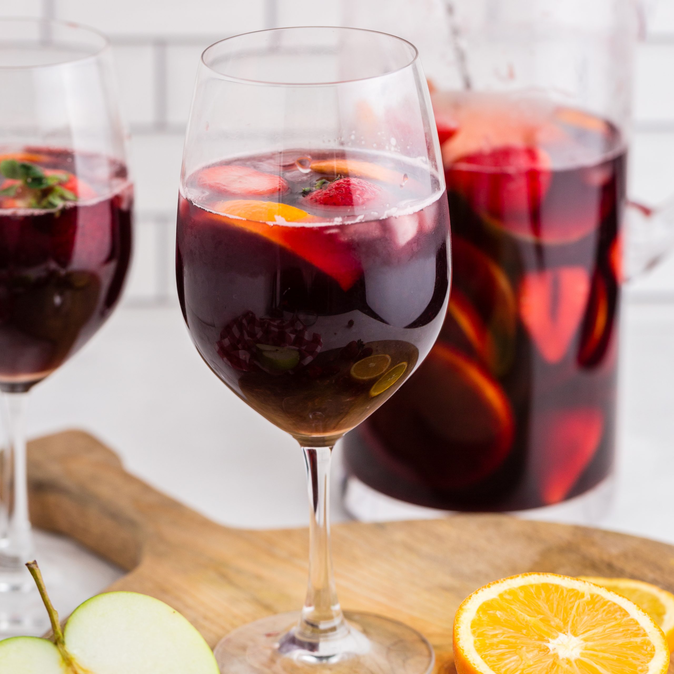 red wine sangria in wine glasses with pitcher in background and sliced apples and oranges on cutting board