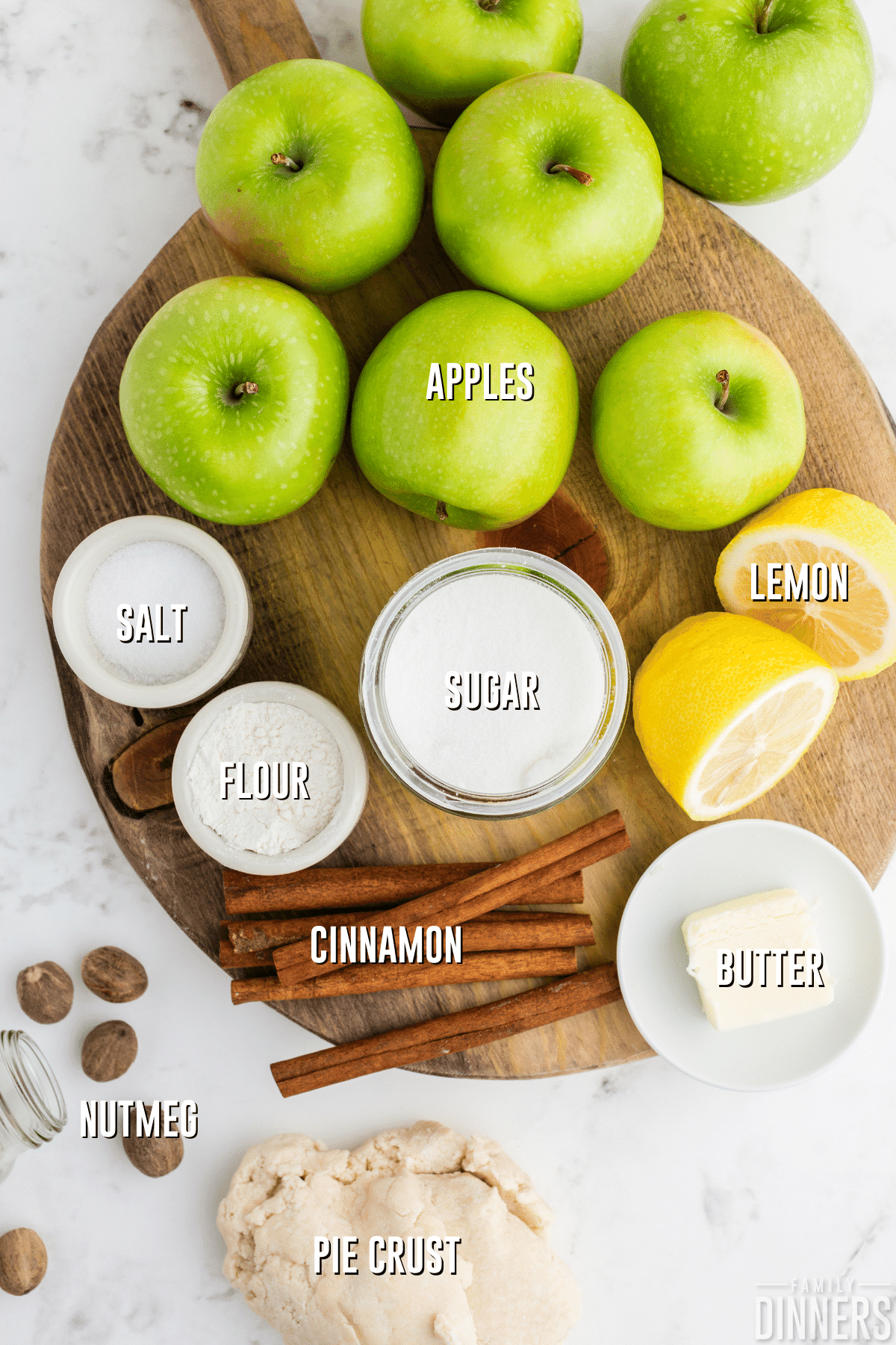 apple pie filling ingredients on a cutting board. 7 green granny smith apples, small bowl of salt, flour, sugar, 5 cinnamon sticks and 5 nutmegs, and a lemon cut in half.