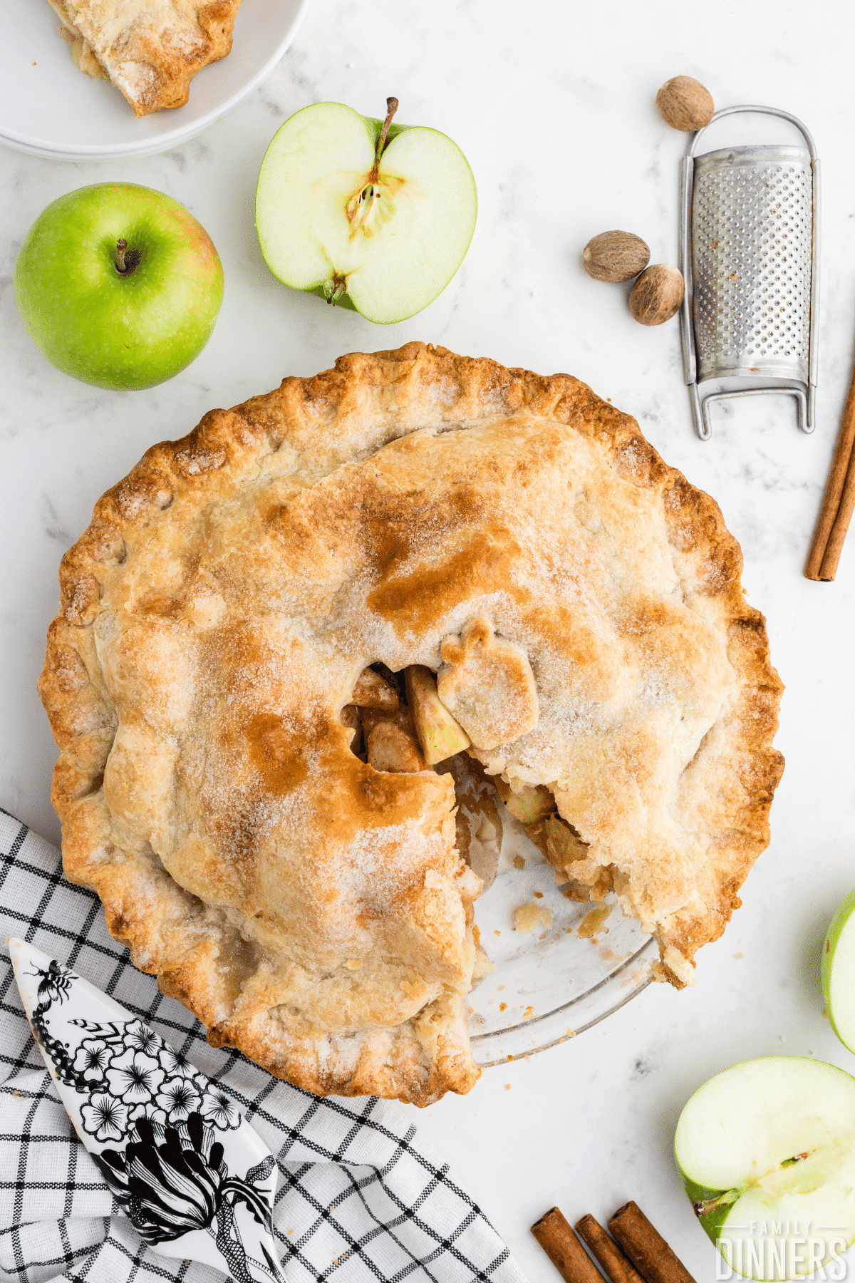granny smith apple pie with a golden pie crust, one slice missing. Counter surrounding pie has 2 cut apples, some nutmegs and cinnamon sticks and a metal tool for shredding the nutmeg.