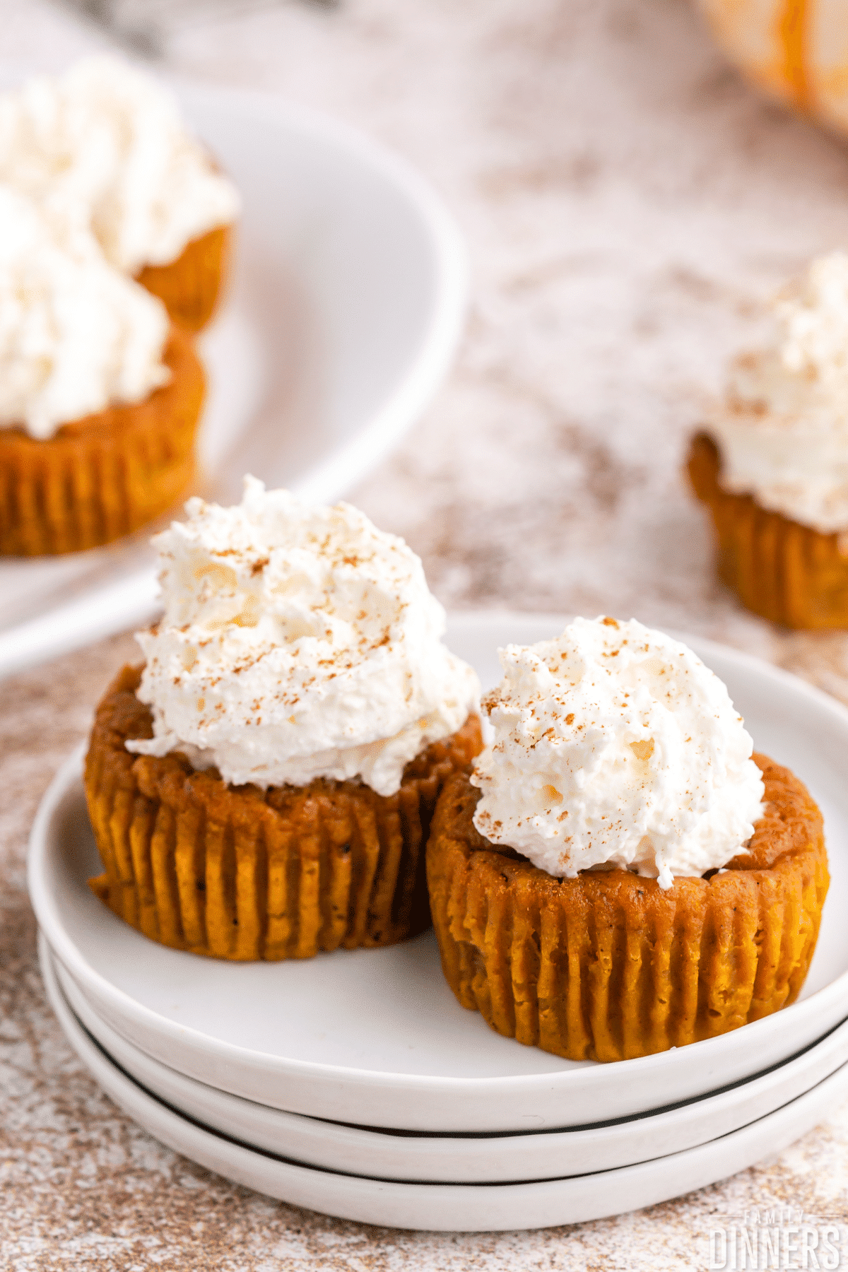2 mini pumpkin pie cupcakes on white plates. This thanksgiving dessert has whipped cream on top and pumpkin pie spice sprinkled on top.