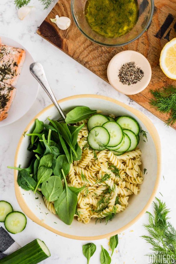fresh spinach, fresh sliced cucumbers and cooked corkscrew pasta in a white bowl with tan ring around the top.