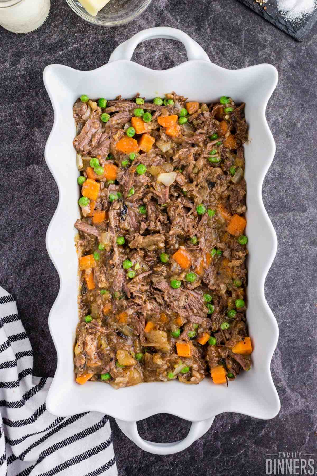 white scalloped casserole dish full of pot roast leftovers shredded beef, carrots and potatoes