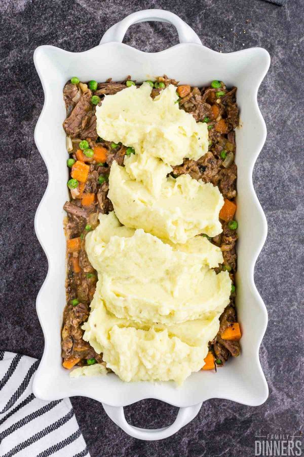 white scalloped dish full of beef, carrots and peas with mashed potato scoops on top