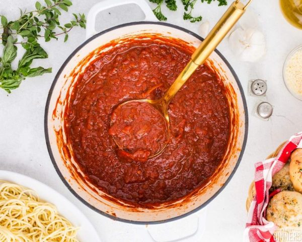white pot of red marinara sauce with gold ladle inside. White marble countertop with basket of rolls and spaghetti next to pot
