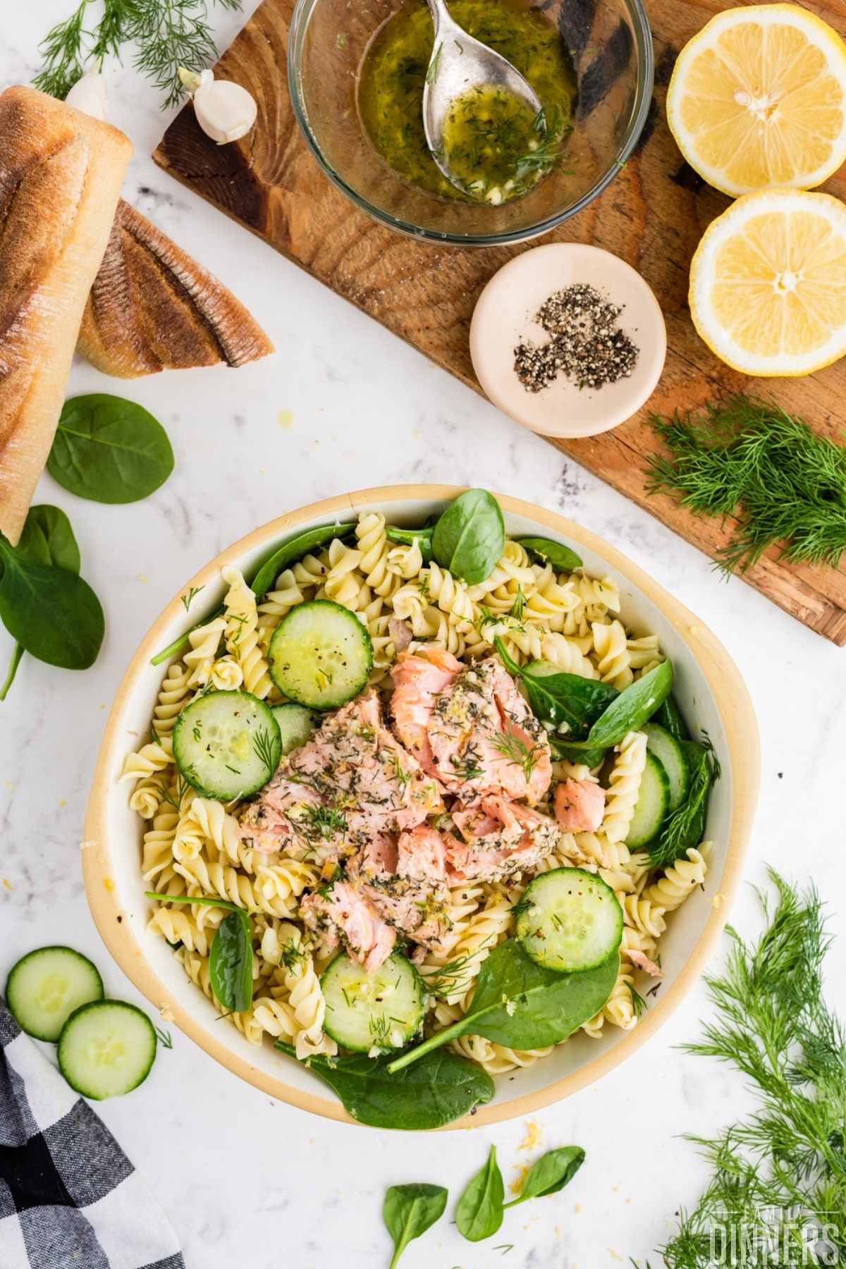 baked salmon leftovers on top of fresh spinach, fresh sliced cucumbers and cooked corkscrew pasta in a white bowl with tan ring around the top.