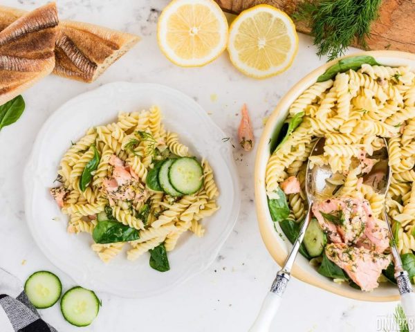 recommended recipe: salmon pasta salad. Image of white marble counter. Large bowl full of pasta, salmon and cucumbers. White plate next to bowl with serving of salmon pasta salad.