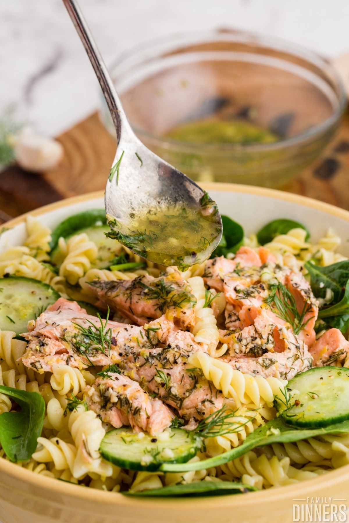 close up of large bowl of salmon pasta with sliced cucumber salad and fresh spinach. Small bowl with lemon vinaigrette next to cold pasta salad. Baked salmon leftovers are on top of the salad.