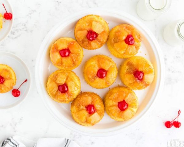 recommended recipe: pineapple upside down mini cakes. Image of white marble counter with round white plate full of 8 mini pineapple upside down cakes with red cherries on top