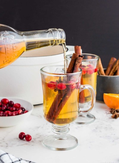 two clear glass mugs filled with golden apple cider cocktail with cranberries, cinnamon sticks in them. Bottle of Tuaca pouring into one mug..