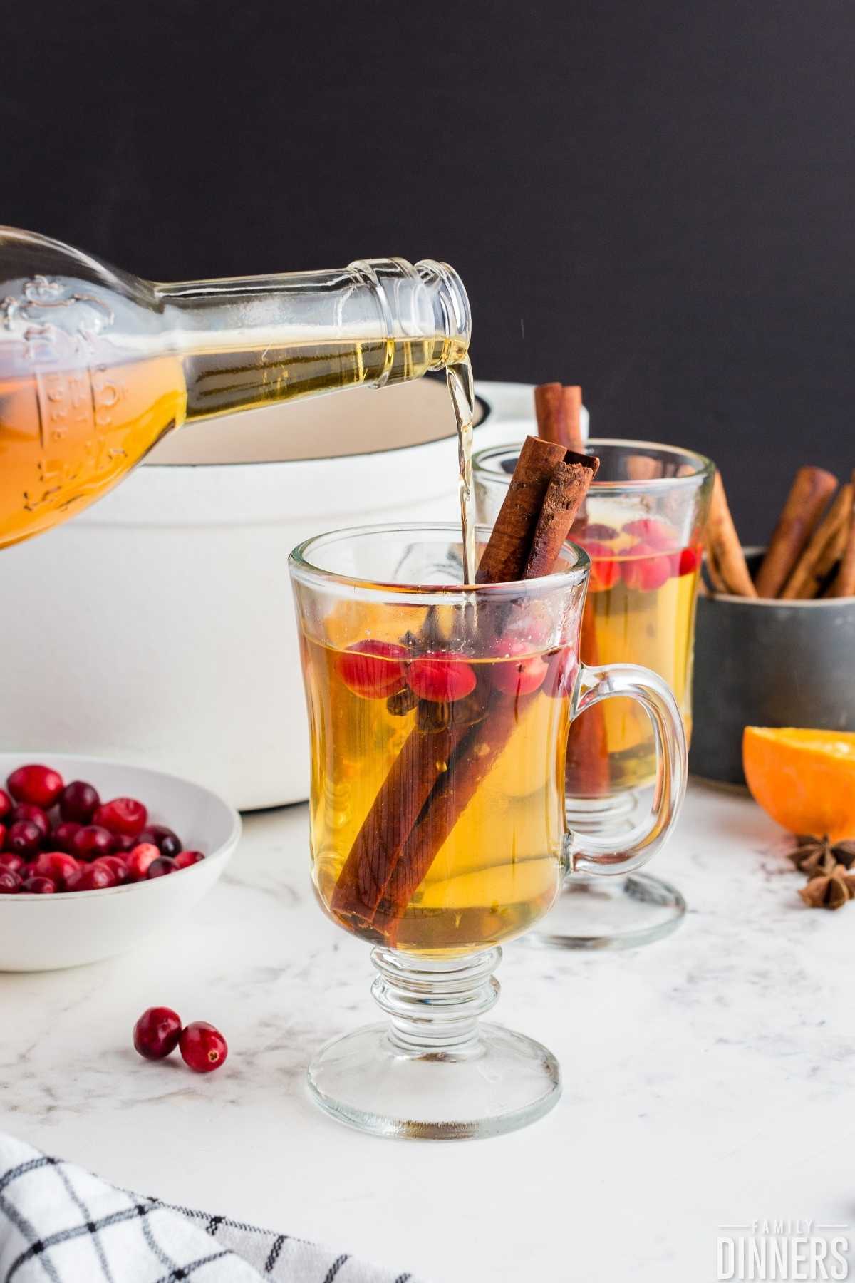 two clear glass mugs filled with golden apple cider cocktail with cranberries, cinnamon sticks in them. Bottle of Tuaca pouring into one mug..