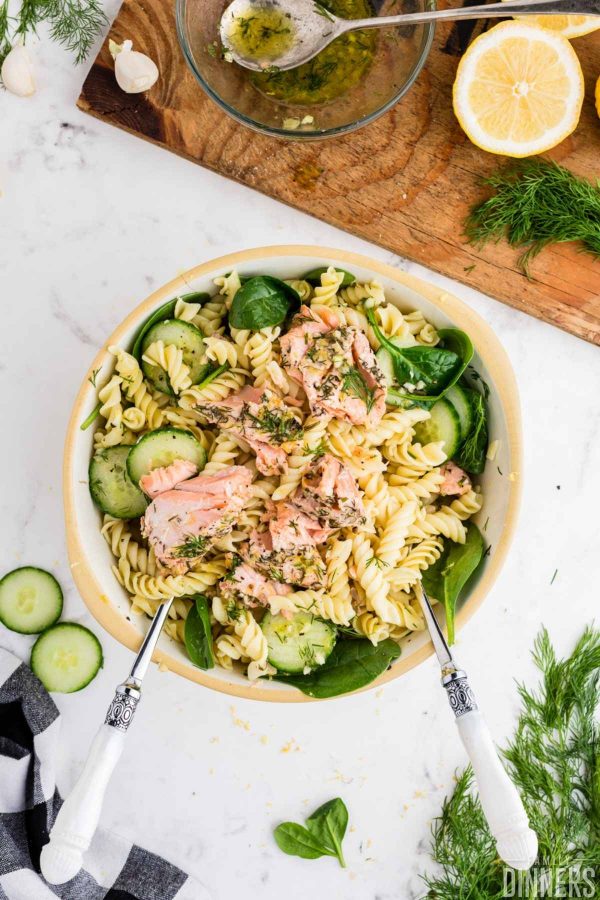 close up of large bowl of salmon pasta salad with sliced cucumber and fresh spinach. Small bowl with lemon vinaigrette next to pasta salad. Two large utensils in the bowl after tossing pasta.