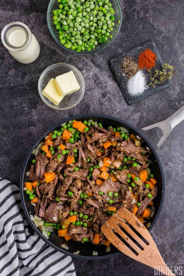 black saute pan with pot roast leftovers, carrots, peas being stirred by a wood slotted spoon