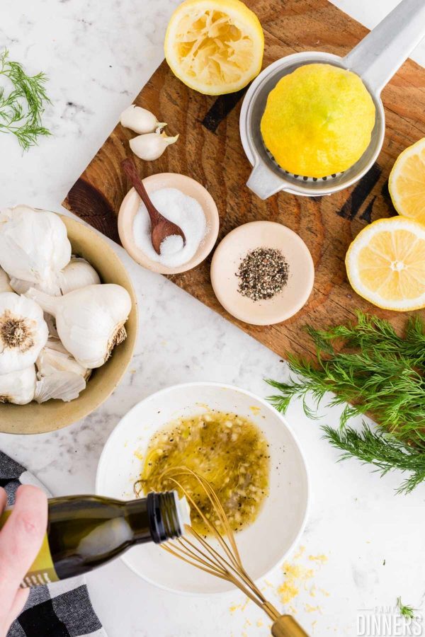 overhead image of lemon dill vinaigrette ingredients including whole garlic in a bowl, sugar in a small bowl, pepper in small bowl, cut lemon, fresh dill and a bowl of ingredients being whisked together.