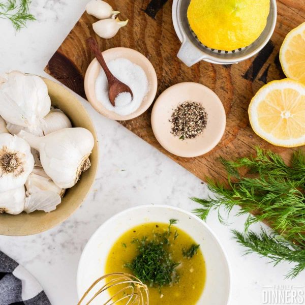 overhead image of lemon dill vinaigrette ingredients including whole garlic in a bowl, sugar in a small bowl, pepper in small bowl, cut lemon, fresh dill and a bowl of ingredients being mixed with gold wisk.
