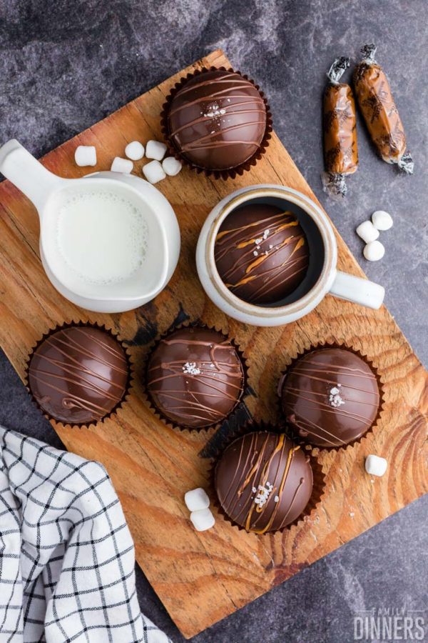 finished hot cocoa bombs on a wood plank on a gray marbled counter. Chocolate bombs are in black cupcake papers and finished with swirls of chocolate and caramel and salt on top of them. Small white pitcher of steaming milk next to the chocolate bombs.