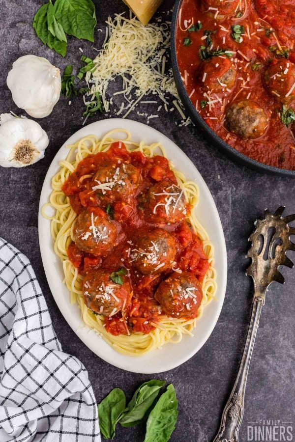 birds eye view of an oval personal serving plate full of cooked spaghetti noodles covered in marinara and 6 meatballs. Serving spoon sits next to plate as well as saute pan with meatballs.