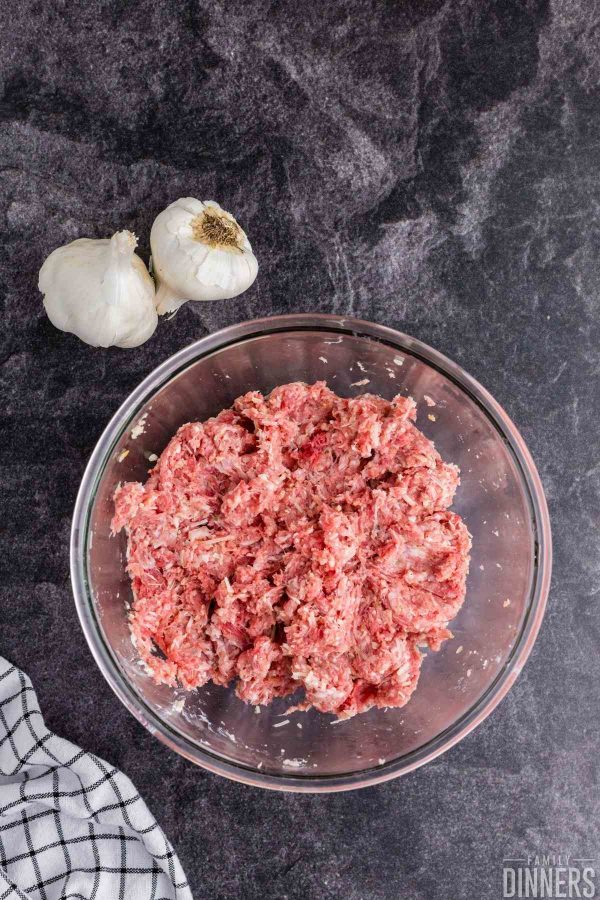 Clear glass bowl on a gray counter. Bowl is full of raw meat. Two whole garlics next to