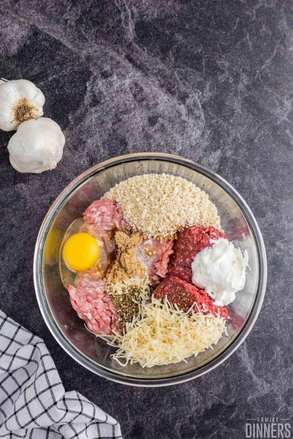 birds eye view of medium sized glass bowl on a dark gray counter. Bowl has raw meat, shredded parmesan cheese, a pile of bread crumbs, a raw egg, yogurt and spices inside it.