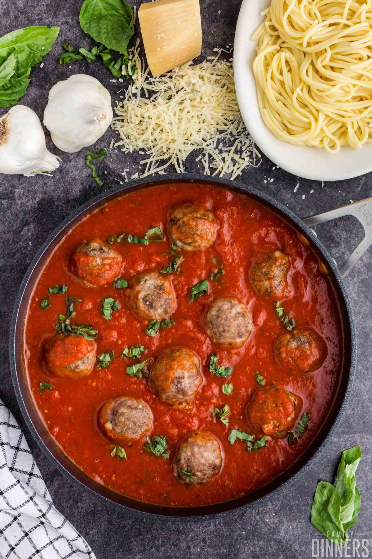 birds eye view of a black large saute pan on a dark gray counter. Pan is full of red marinara sauce, large cooked meatballs and sprinkled with fresh slices of basil.