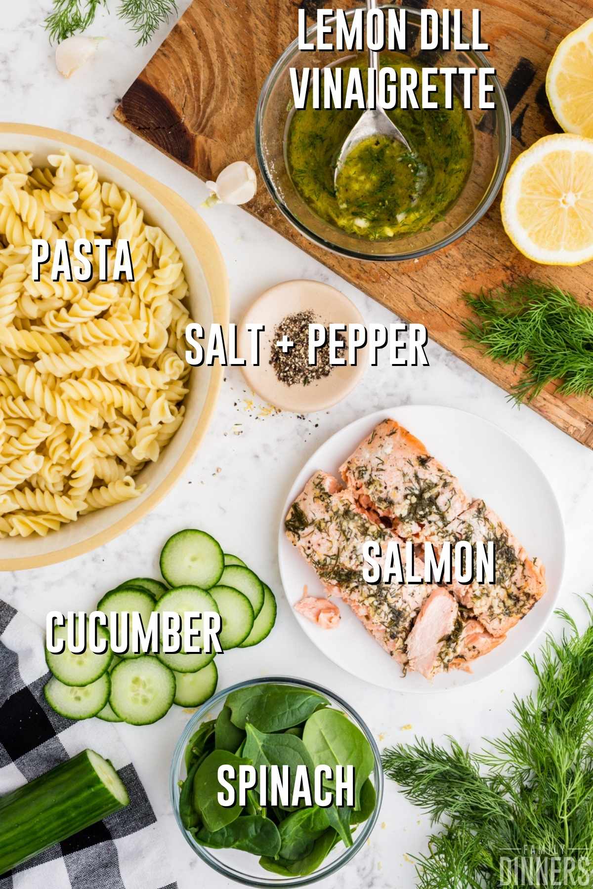  ingredients - white counter with bowl of corkscrew pasta cooked, sliced cucumbers, small bowl of fresh spinach, round white plate of lemon dill salmon, salt and pepper, bowl of lemon dill vinaigrette, sliced lemons and fresh dill. The salmon pasta spinach cucumber salad ingredients are all fresh and bright.