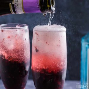 champagne being poured into two champagne glasses full of blackberry puree in bottom of glass. Bubbles foaming out of top of glass
