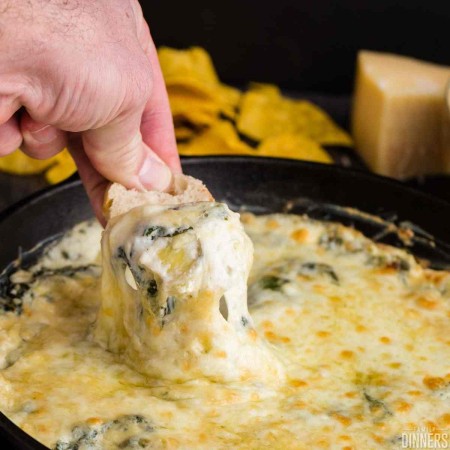 crusty bread being dipped into hot cheesy spinach and artichoke dip in a black cast iron pan