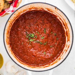Savory looking marinara sauce with red wine in a white dutch oven pot garnished with fresh herbs.