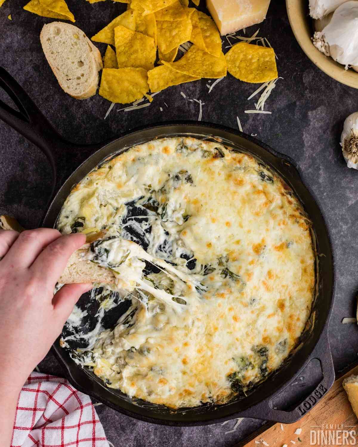 birds eye view of hand dipping bread in cheesy hot spinach dip pan.