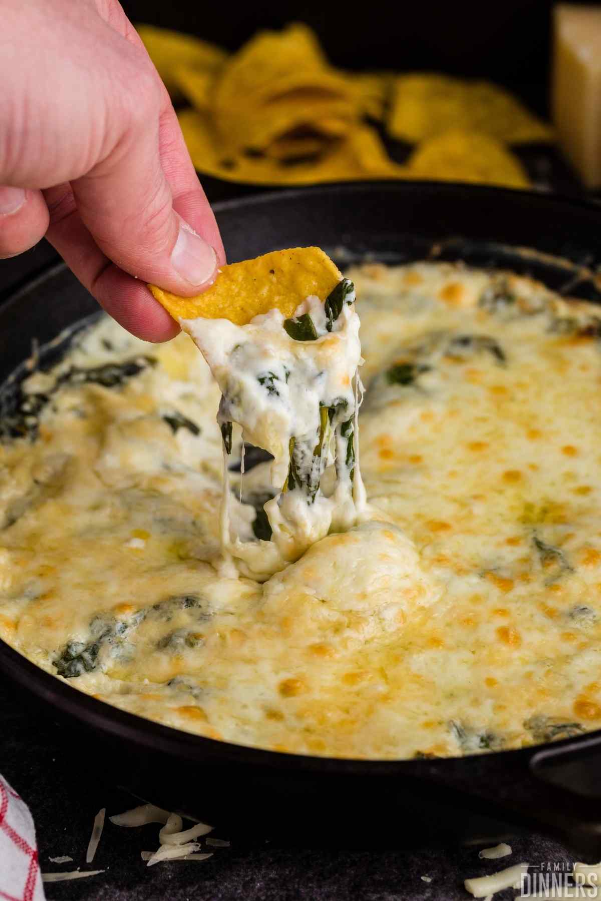 yellow corn chip being dipped into hot cheesy spinach and artichoke dip in a black cast iron pan