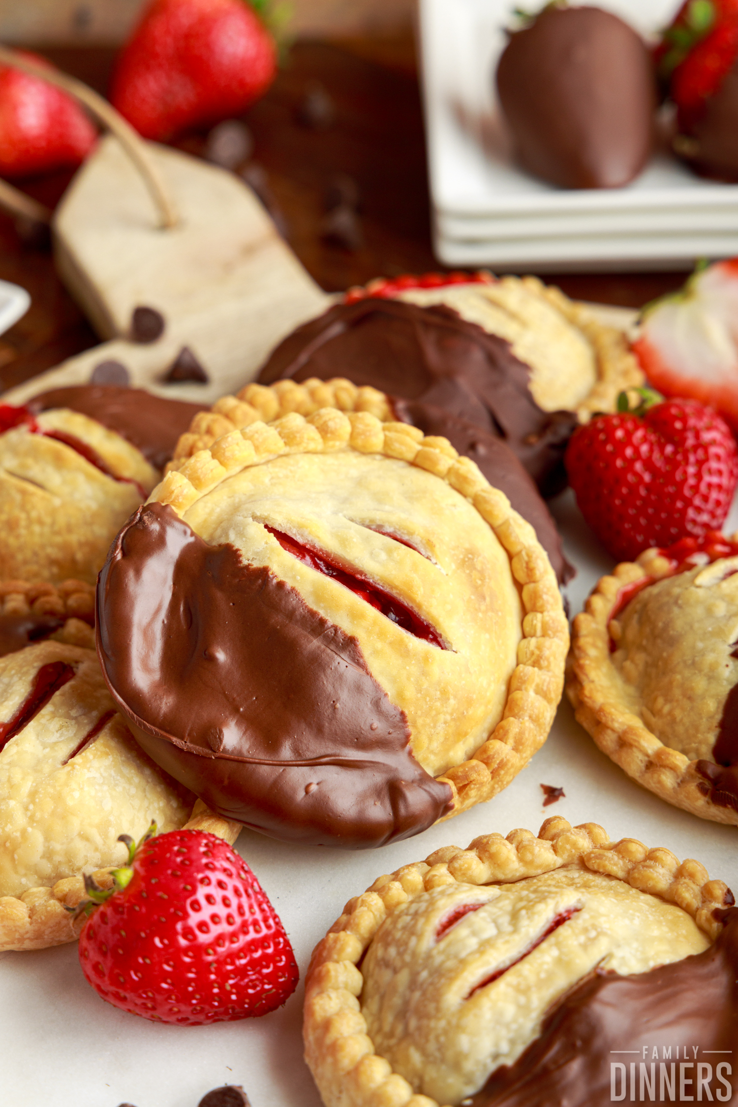a batch of chocolate dipped strawberry hand pies with slits on top next to whole berries