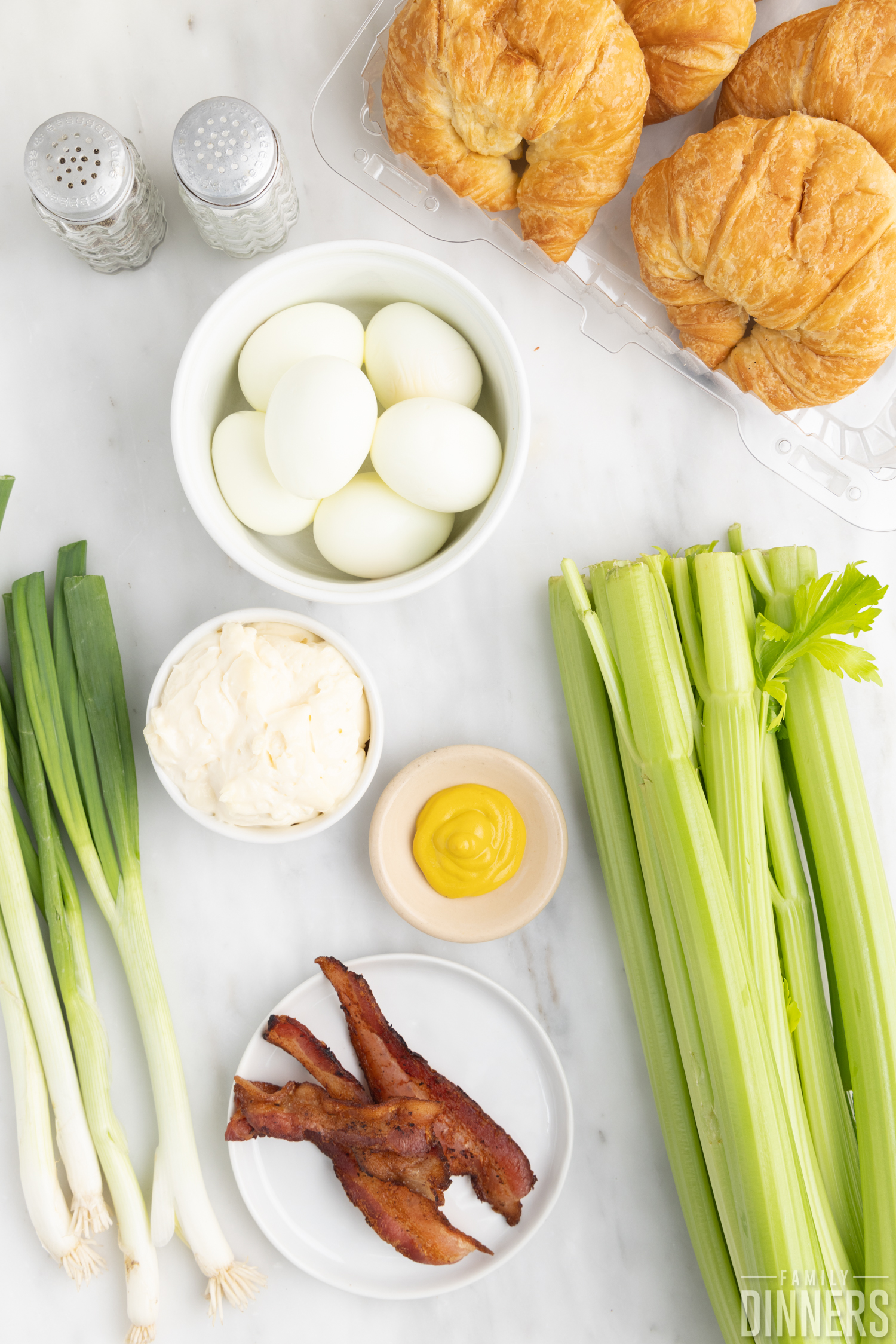 Ingredients for egg salad sandwich on countertop--eggs, salt and pepper, green onions, celery, eggs and mustard, and croissants.