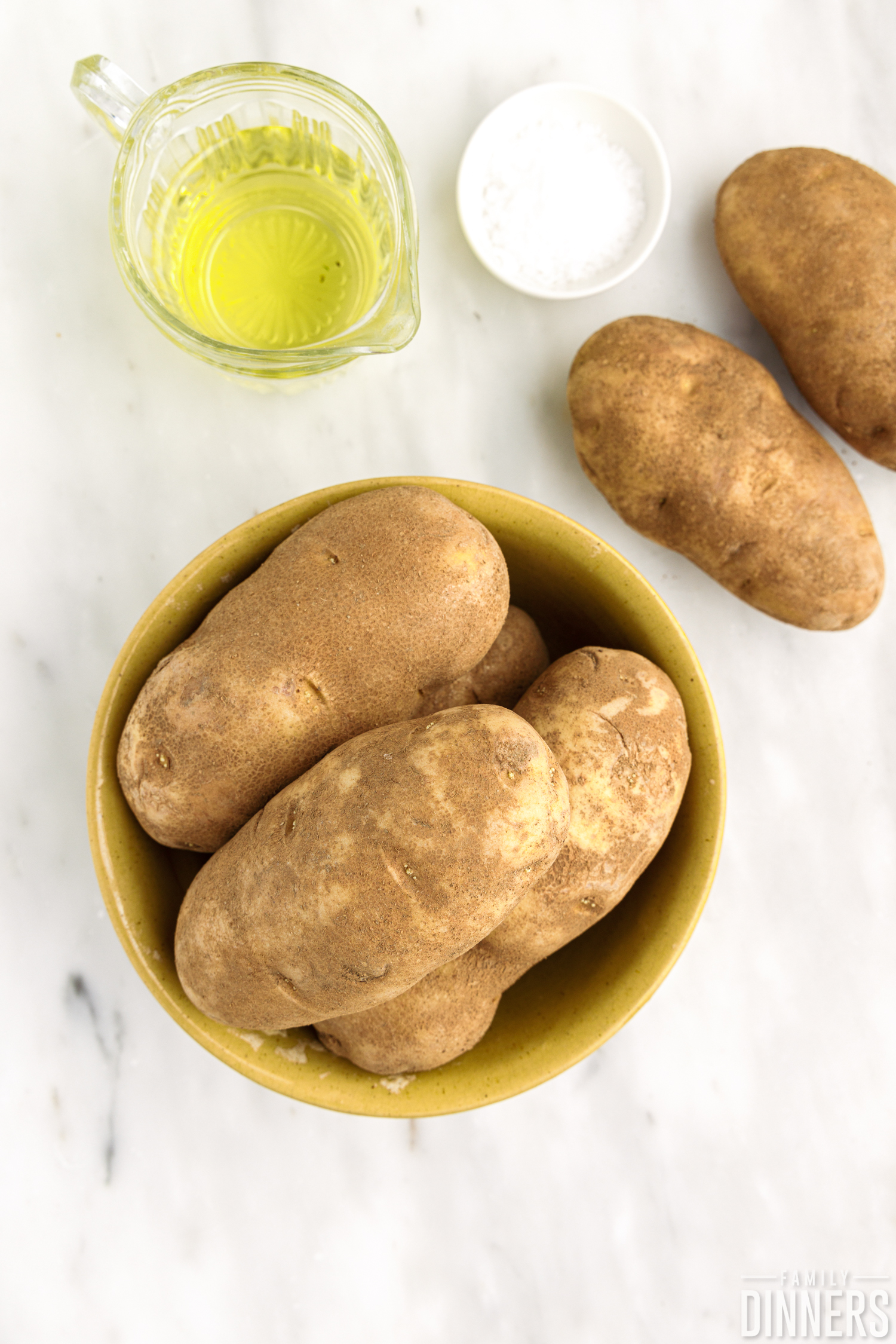 Raw Russet potatoes on marble countertop next to a small pitcher of oil and small bowl of coarse salt.