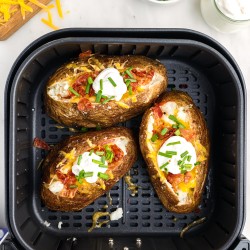 Baked potatoes in air fryer basket loaded with toppings like cheddar cheese, bacon bits, sour cream and green onion pieces.
