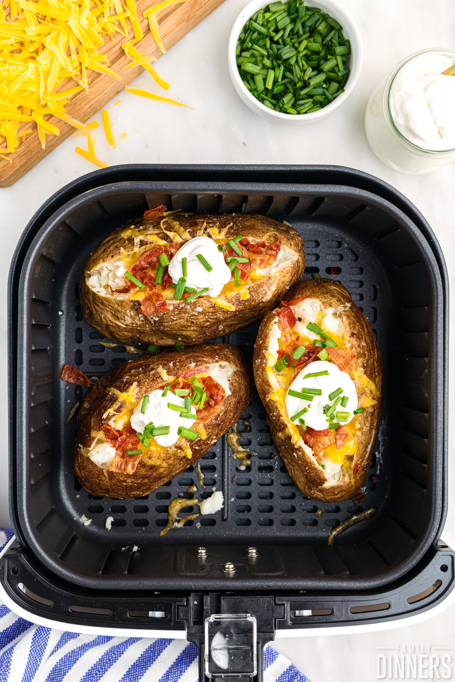 Close up of baked potatoes in an air fryer basket, fully loaded with sour cream, cheddar cheese, bacon and green onions.