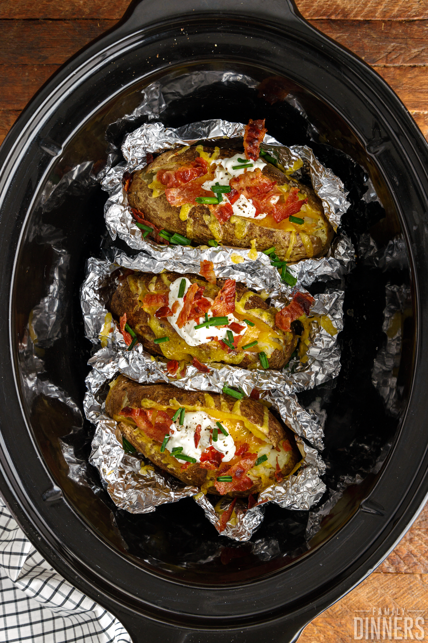 Fully-cooked baked potatoes in open foil inside a crock pot, loaded with cheddar cheese, sour cream, bacon bits and chives.