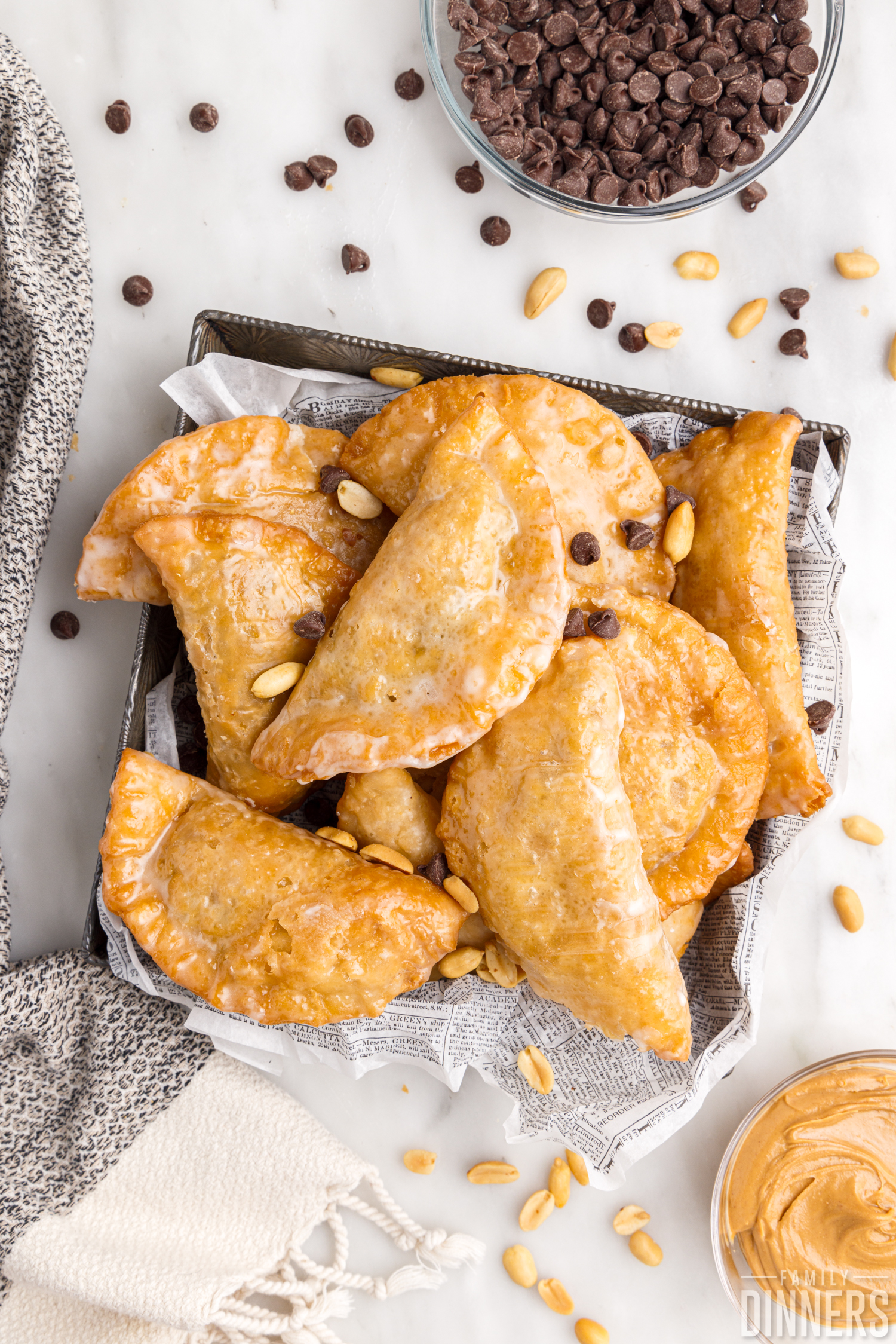 pile of deep fried hand pies sitting in a square tin, sprinkled with chocolate chips and peanuts, a bowl of chocolate chips and a bowl of peanut butter are sitting next to them 