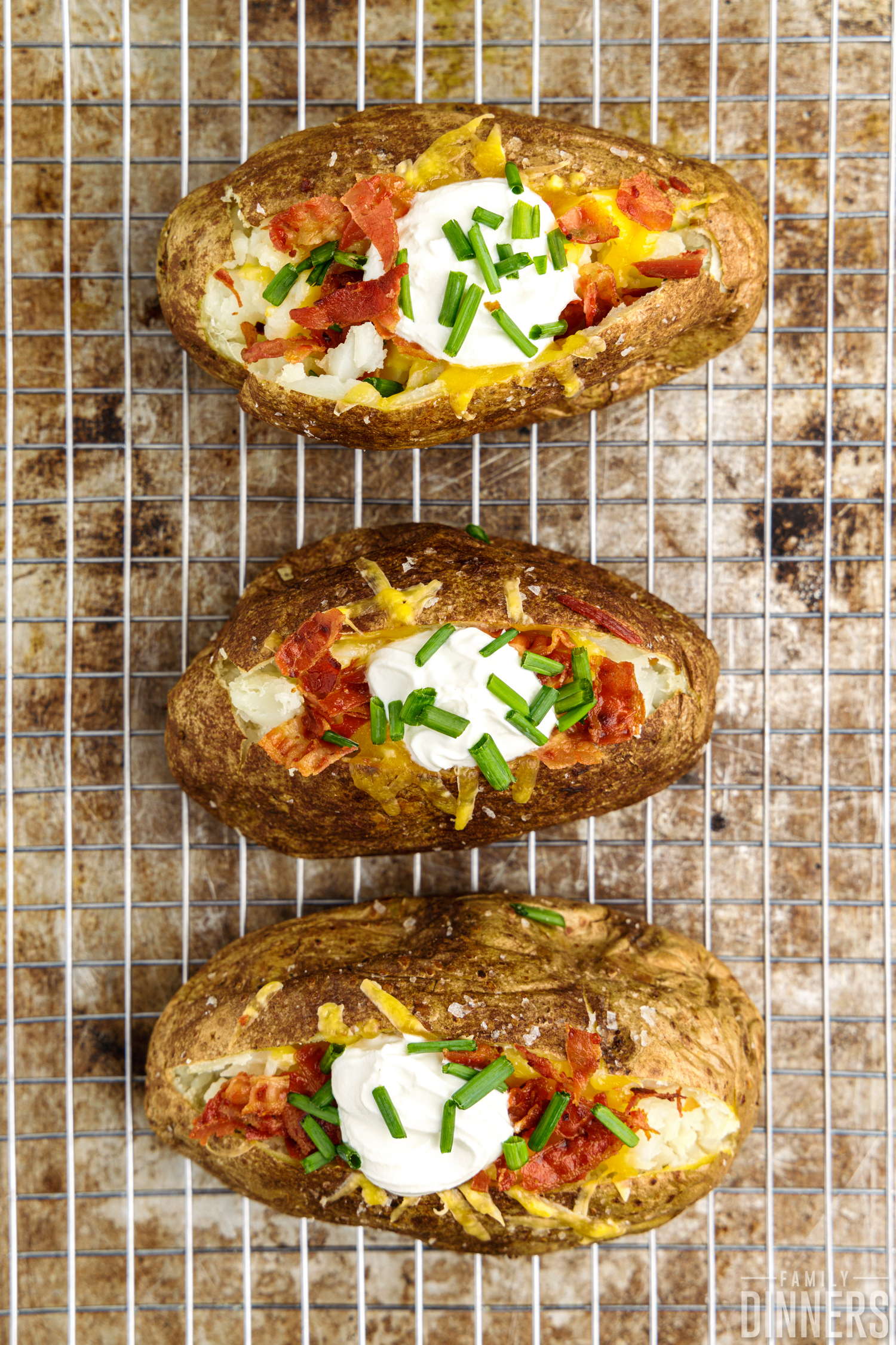 Fully-cooked baked potatoes on wire rack cut open and topped with cheese, sour cream, bacon and green onions.