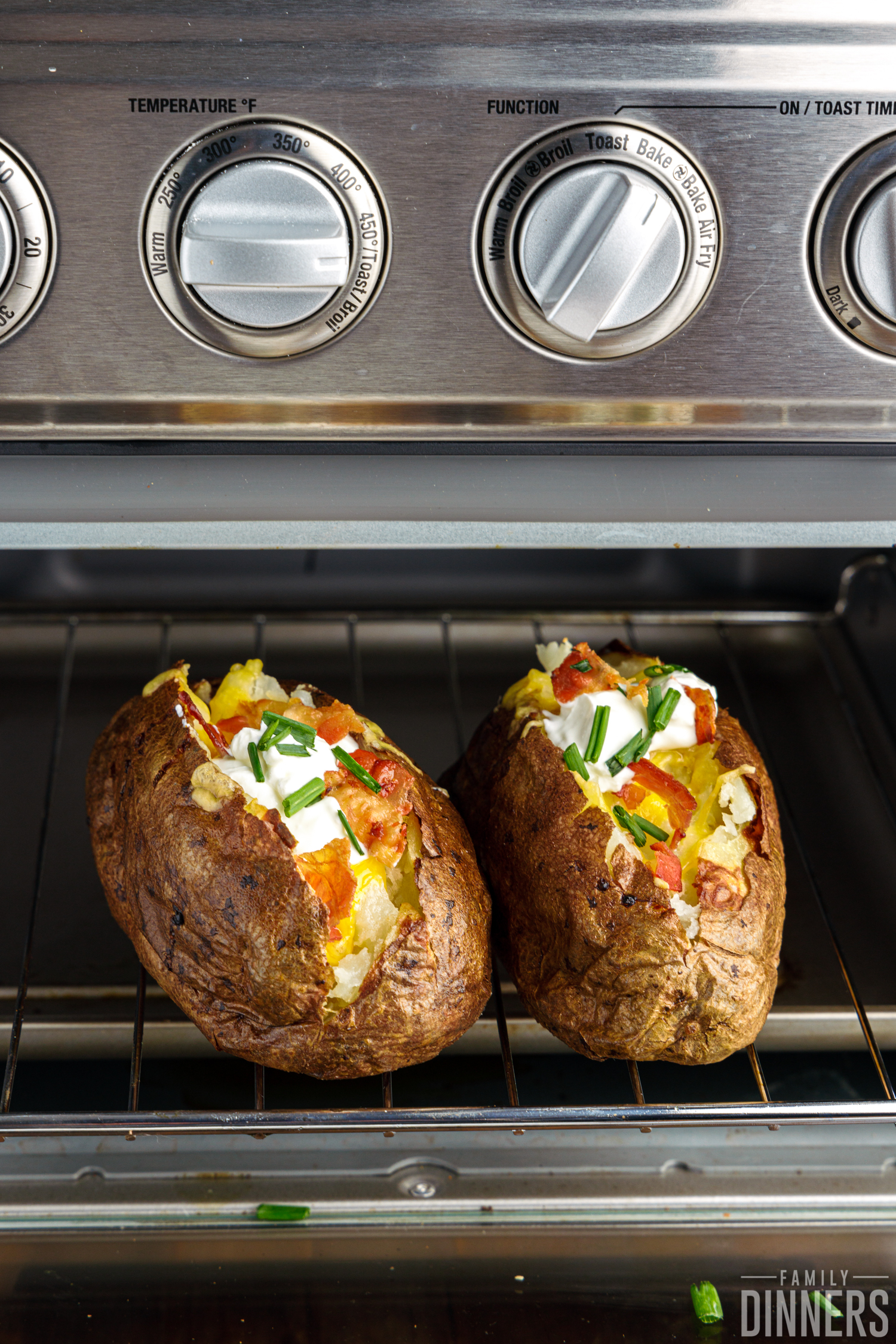 Two fully loaded potatoes in toaster oven topped with cheese, bacon its, sour cream, and chives.