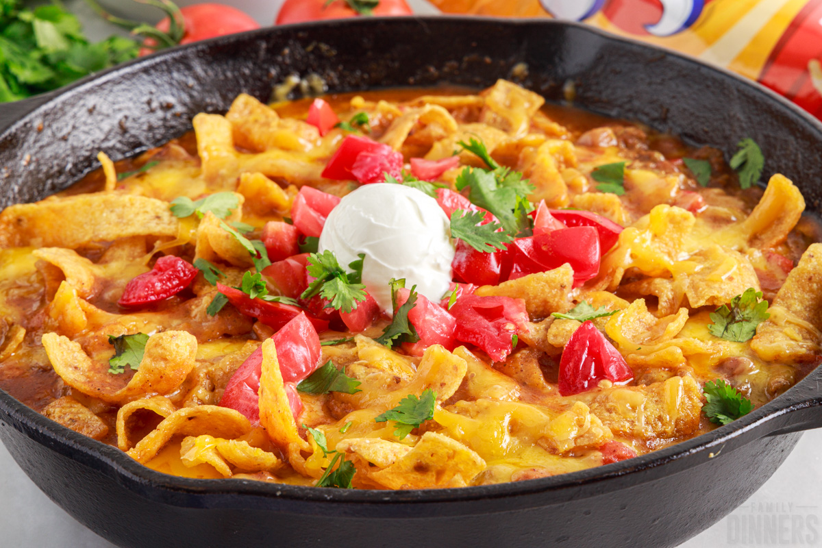 ready to serve Frito pie in a skillet topped with diced tomatoes, cilantro and a dollop of sour cream