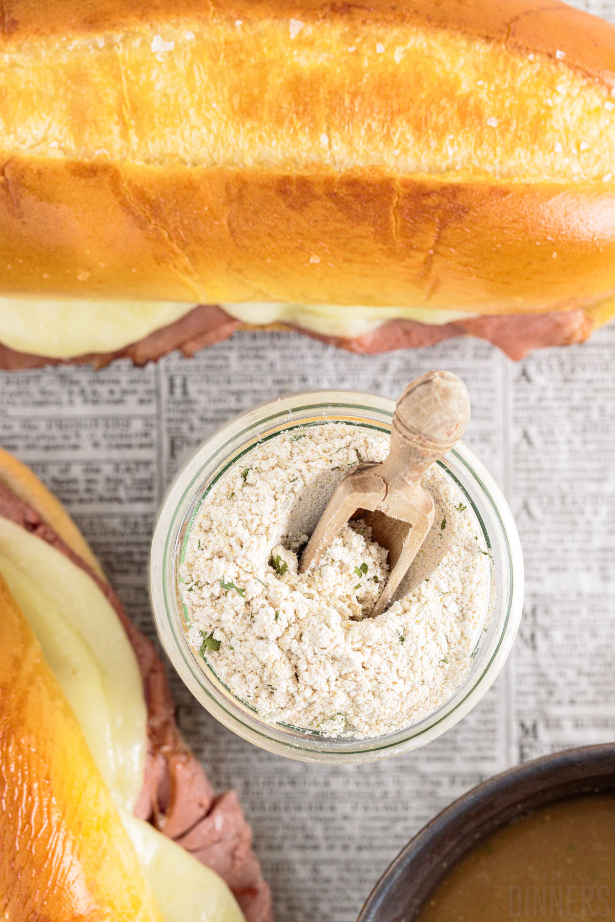 dry au jus seasoning mix in a glass jar with a wooden spoon with French dip sandwiches